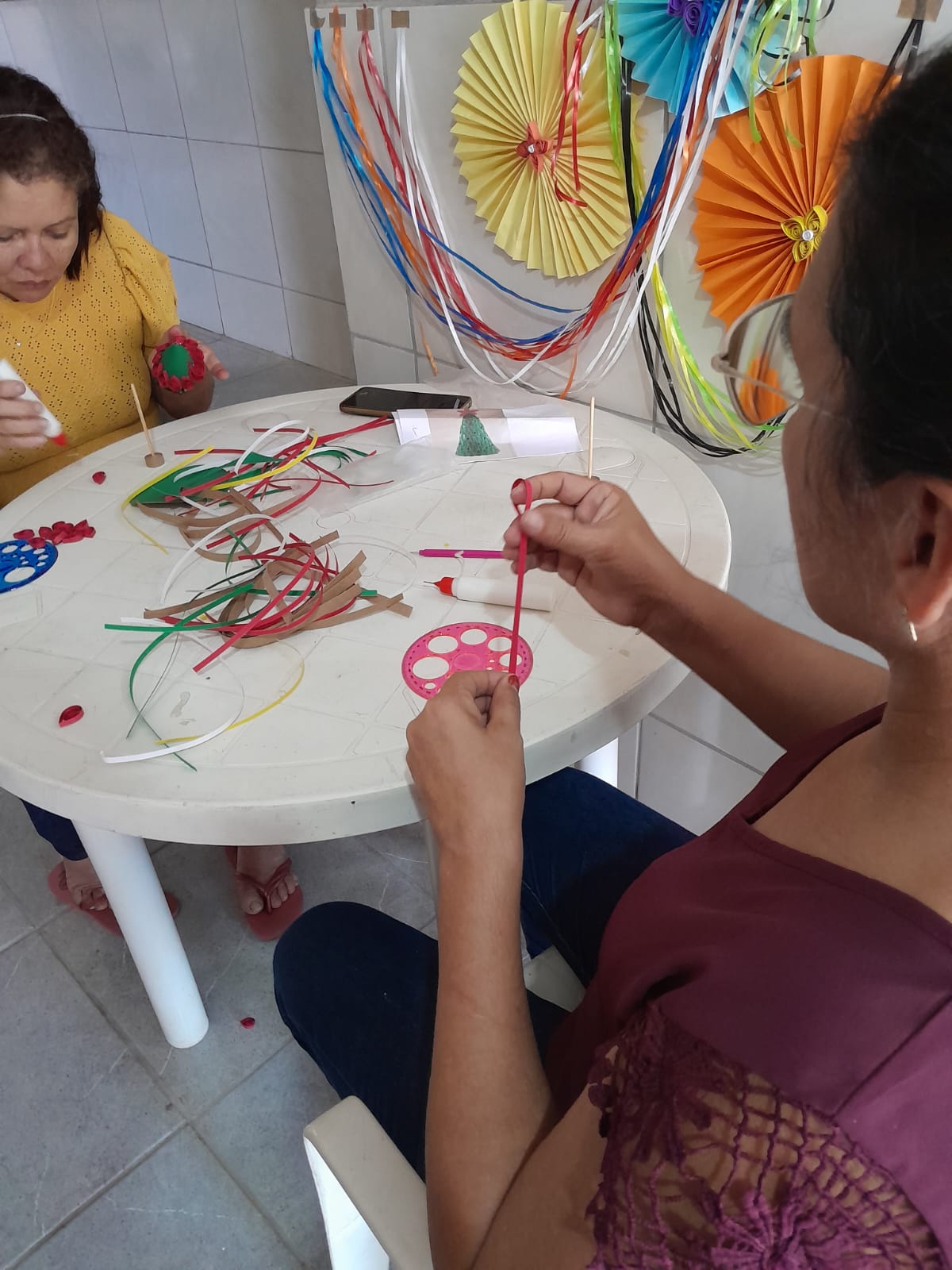 Duas mulheres trabalhando com fitas coloridas e enfeites de papel em mesa redonda, com leques decorativos na parede.