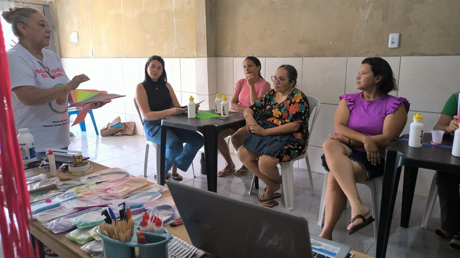 Instrutora apresenta papéis e materiais coloridos para o grupo na oficina de quilling e artesanato.