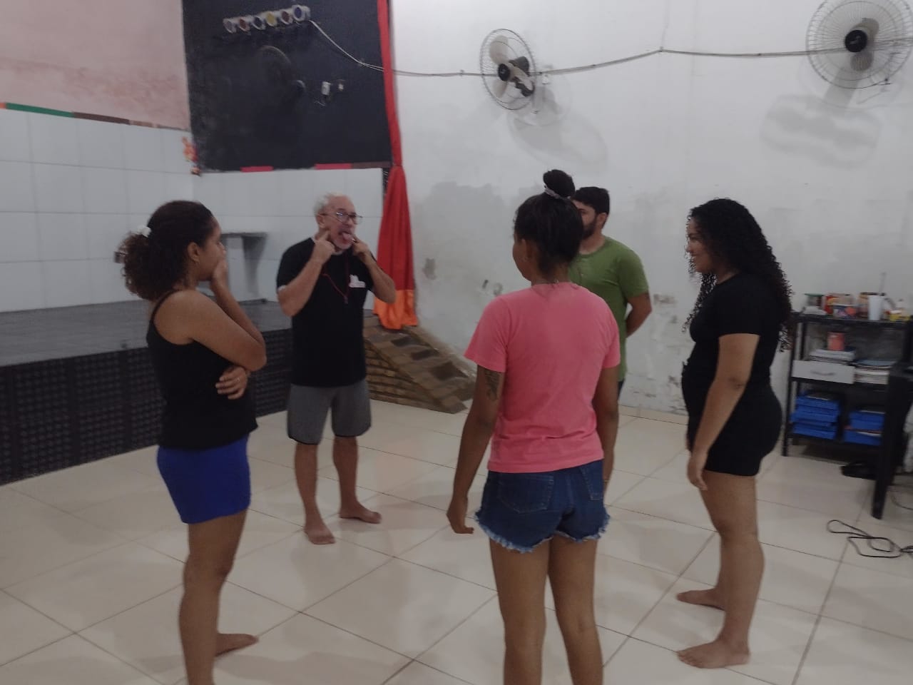 Cinco pessoas descalças em círculo no salão da Fundação, com o instrutor conduzindo um exercício de teatro.